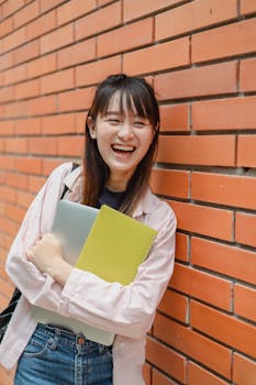Happy young woman student with laptop, smiling and leaning against a brick wall outdoors.